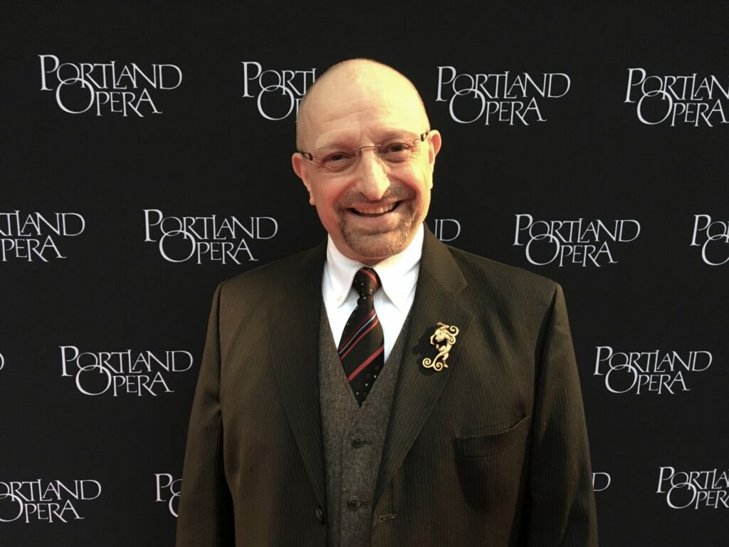 A man smiles while wearing a suit and standing in front of a background that says "Portland Opera."