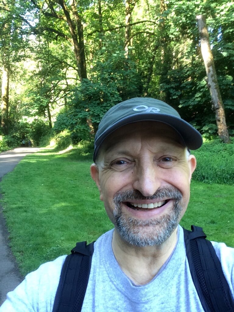 A man wearing a cap smiles at the viewer. He is in nature.